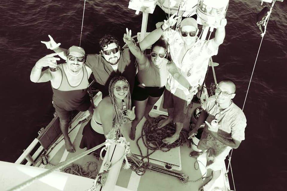 A group of six friends smiling and posing on the deck of a sailboat in Zihuatanejo, Mexico. The photo is taken from above, showing the relaxed, joyful vibe of the group as they enjoy a day out at sea. Everyone is barefoot, wearing casual summer clothes and sunglasses, with the ocean visible all around the boat.